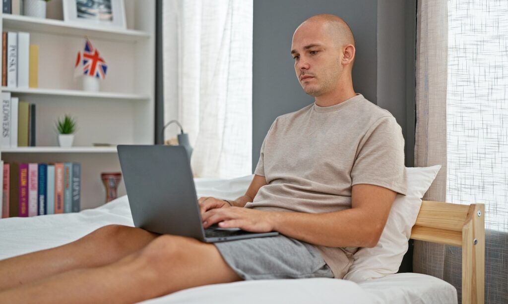 Young hispanic man using laptop sitting on bed at bedroom